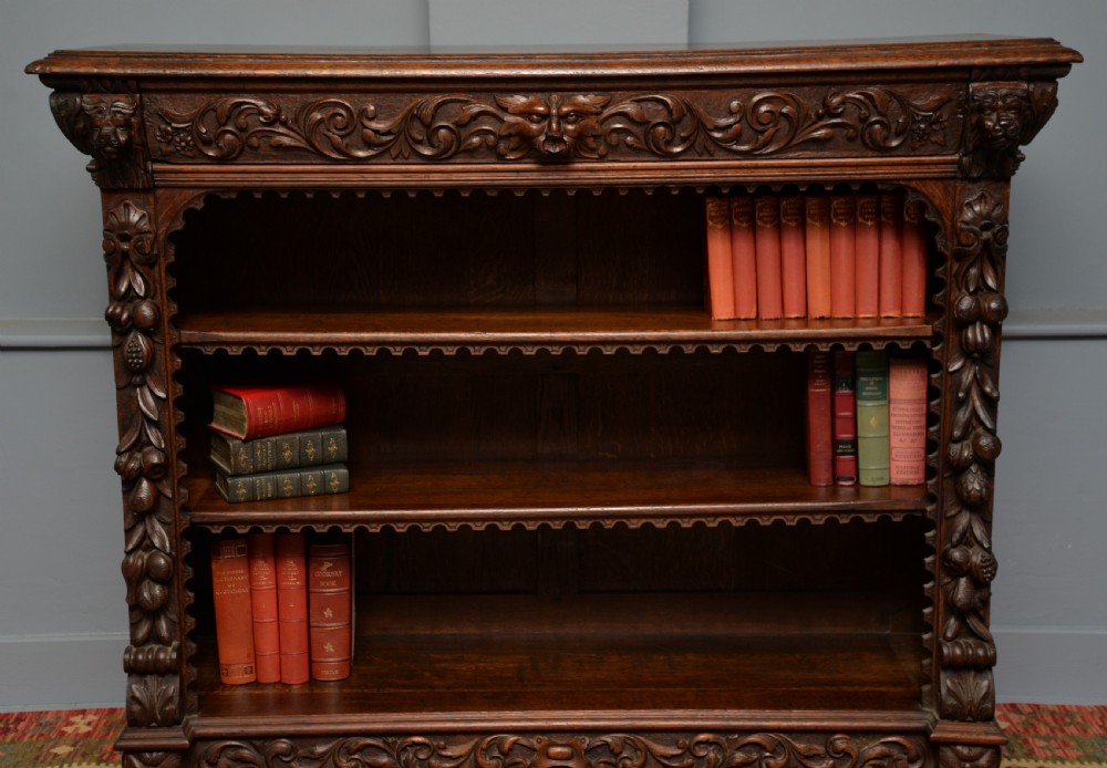 A low solid oak open bookcase decorated with hand-carved Green Man motifs, foliate pilasters, three shelves with scalloped edges, and a plinth base. View II