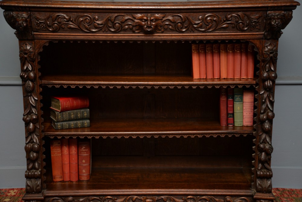 A low solid oak open bookcase decorated with hand-carved Green Man motifs, foliate pilasters, three shelves with scalloped edges, and a plinth base. View III