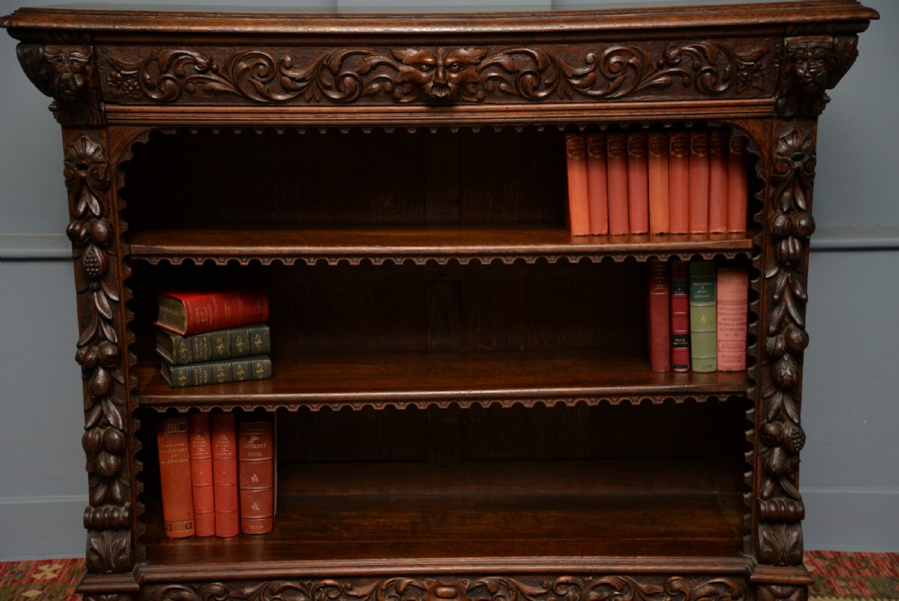 A low solid oak open bookcase decorated with hand-carved Green Man motifs, foliate pilasters, three shelves with scalloped edges, and a plinth base. View V