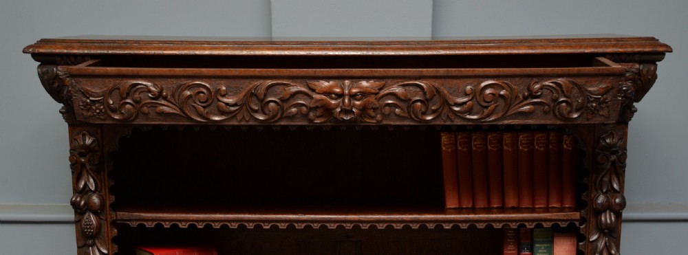 A low solid oak open bookcase decorated with hand-carved Green Man motifs, foliate pilasters, three shelves with scalloped edges, and a plinth base. View VI