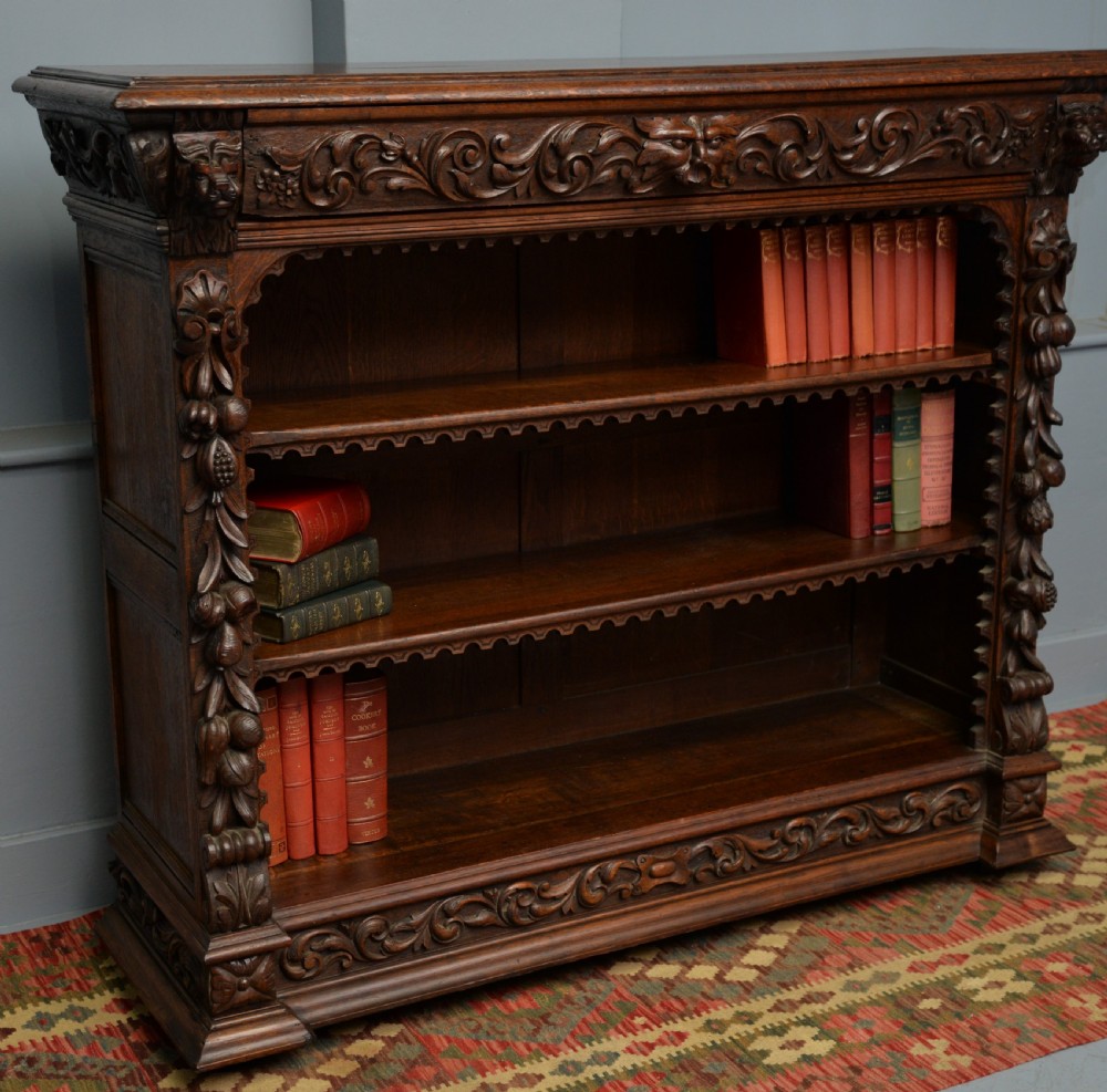 A low solid oak open bookcase decorated with hand-carved Green Man motifs, foliate pilasters, three shelves with scalloped edges, and a plinth base. View VII