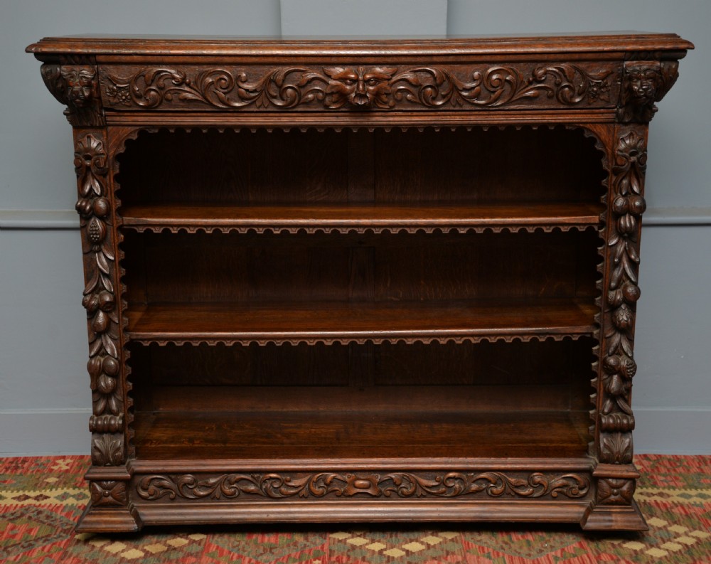A low solid oak open bookcase decorated with hand-carved Green Man motifs, foliate pilasters, three shelves with scalloped edges, and a plinth base. View X