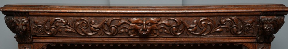 A low solid oak open bookcase decorated with hand-carved Green Man motifs, foliate pilasters, three shelves with scalloped edges, and a plinth base. View XI