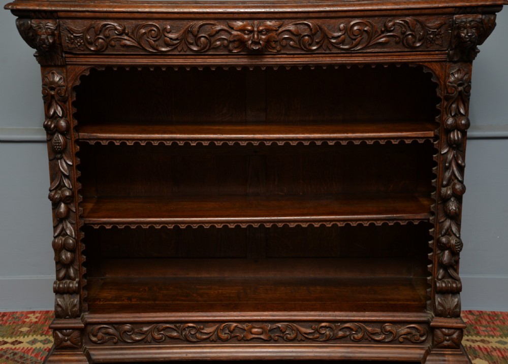 A low solid oak open bookcase decorated with hand-carved Green Man motifs, foliate pilasters, three shelves with scalloped edges, and a plinth base. View XII