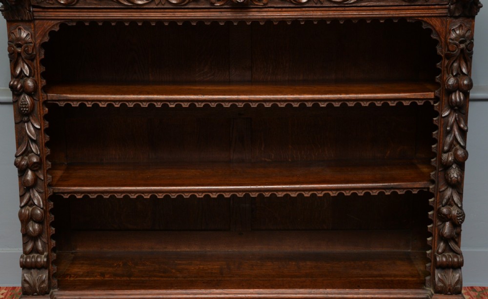 A low solid oak open bookcase decorated with hand-carved Green Man motifs, foliate pilasters, three shelves with scalloped edges, and a plinth base. View XIII