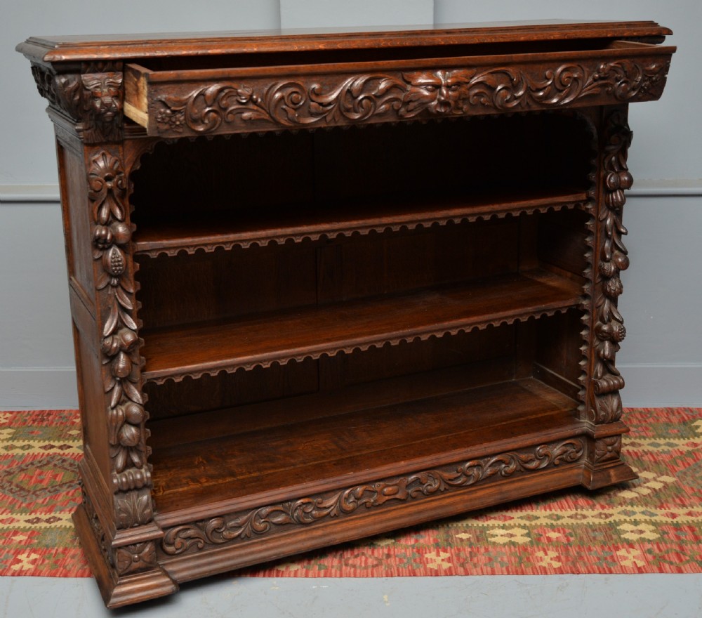 A low solid oak open bookcase decorated with hand-carved Green Man motifs, foliate pilasters, three shelves with scalloped edges, and a plinth base. View XIV