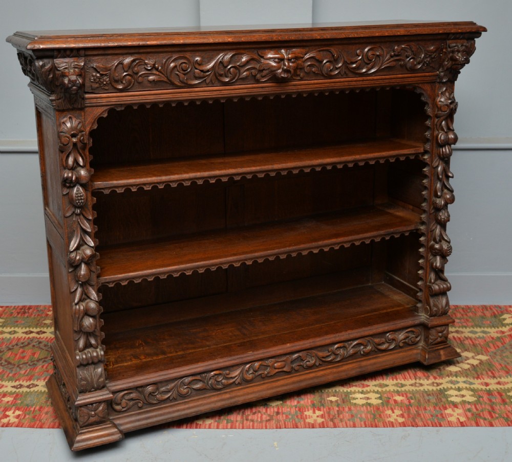 A low solid oak open bookcase decorated with hand-carved Green Man motifs, foliate pilasters, three shelves with scalloped edges, and a plinth base. View XV