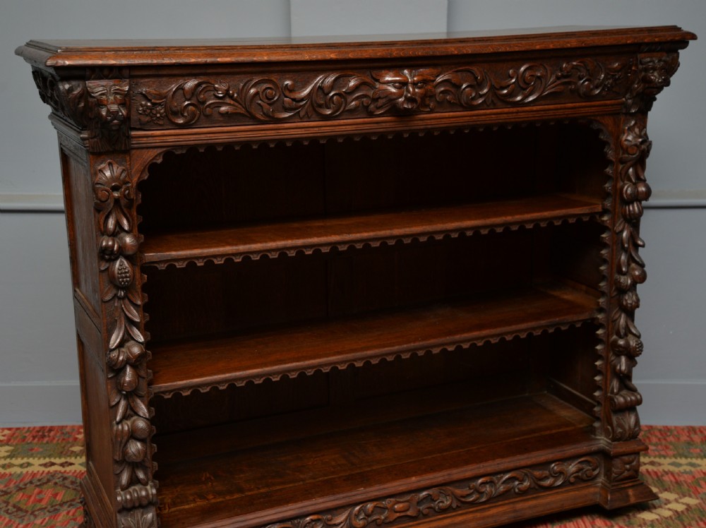 A low solid oak open bookcase decorated with hand-carved Green Man motifs, foliate pilasters, three shelves with scalloped edges, and a plinth base. View XVI