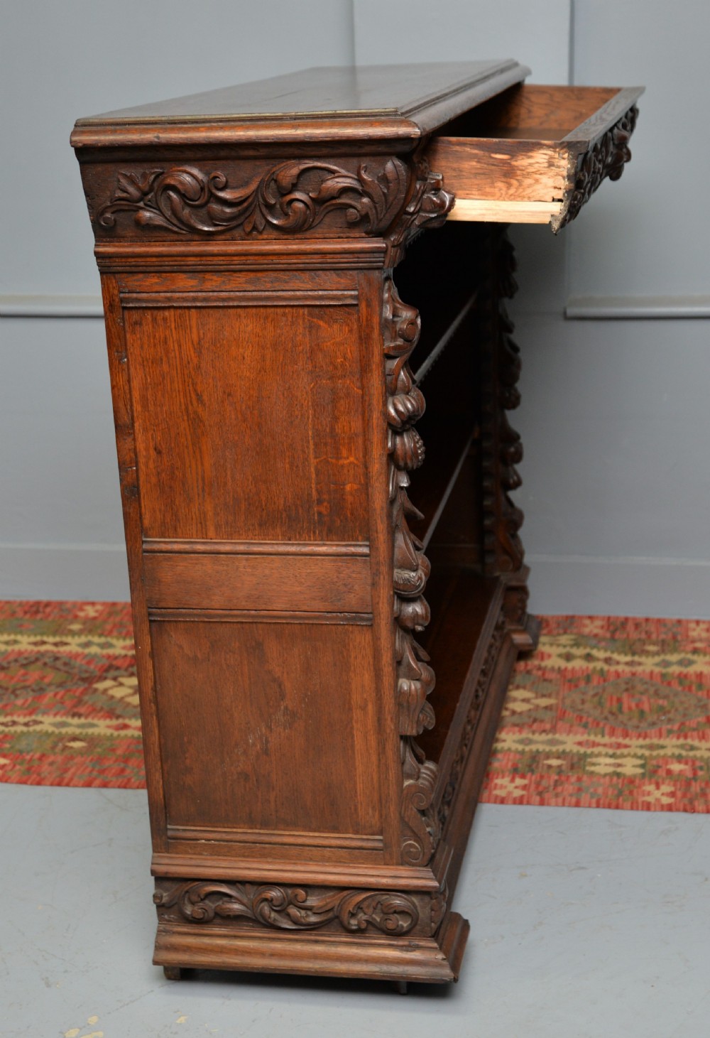 A low solid oak open bookcase decorated with hand-carved Green Man motifs, foliate pilasters, three shelves with scalloped edges, and a plinth base. View XVIII
