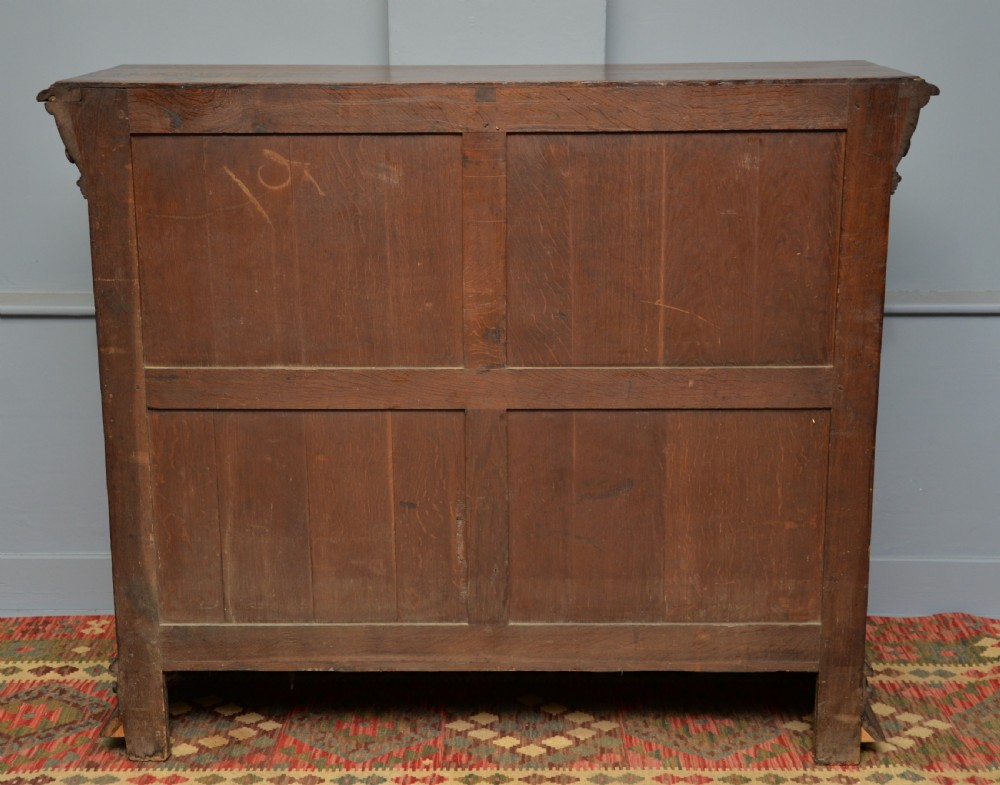 A low solid oak open bookcase decorated with hand-carved Green Man motifs, foliate pilasters, three shelves with scalloped edges, and a plinth base. View XX