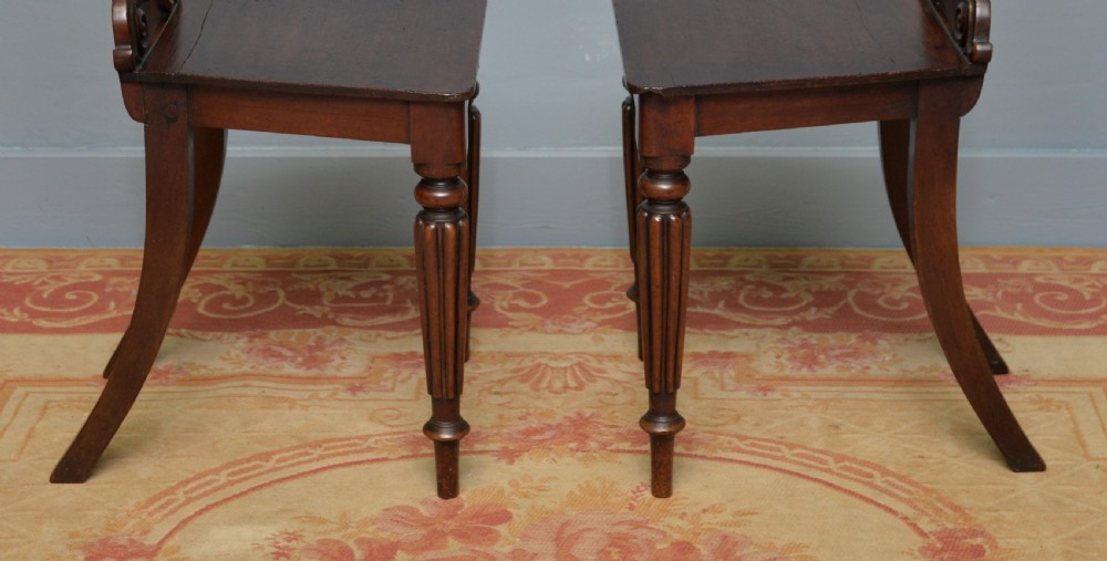 A pair of Regency mahogany hall chairs featuring carved scroll backs with shield motifs and turned reeded front legs. View X