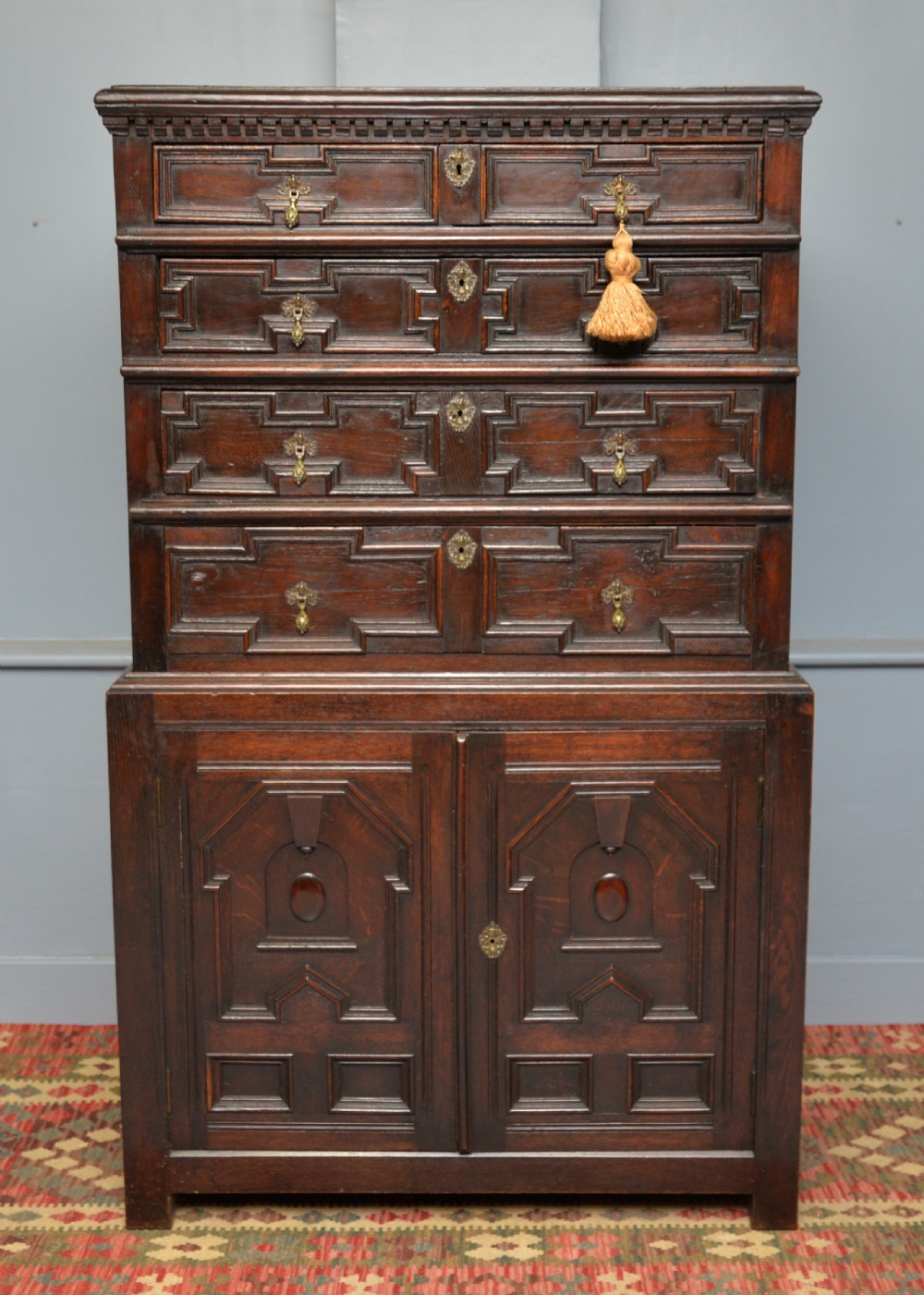 A tall dark oak Charles II chest featuring geometric moulded drawers and brass tear-drop handles above a two-door cupboard.