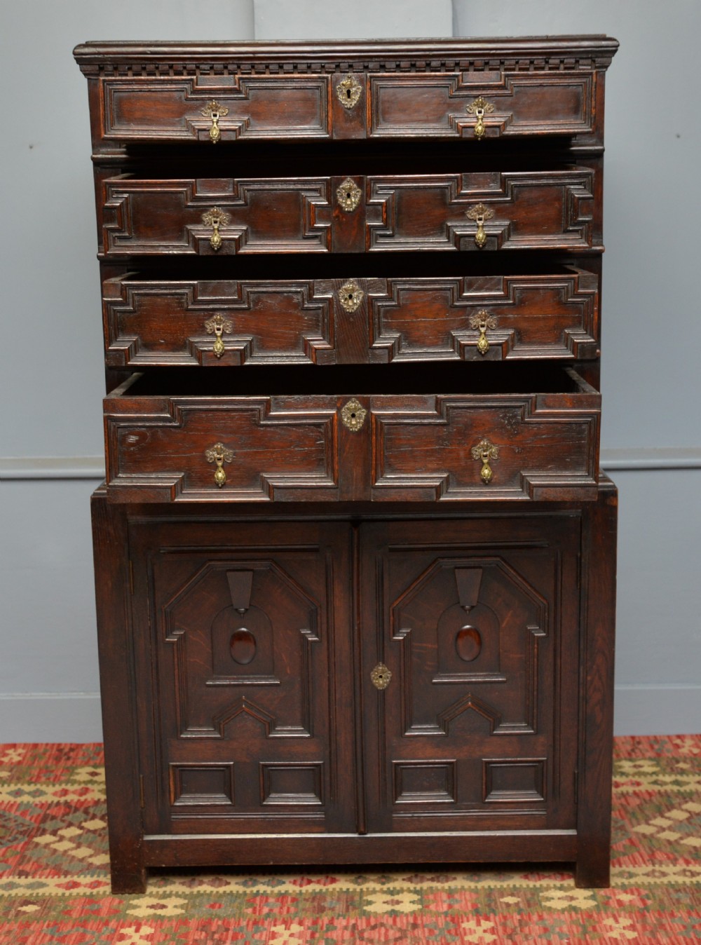 A tall dark oak Charles II chest featuring geometric moulded drawers and brass tear-drop handles above a two-door cupboard. View IX