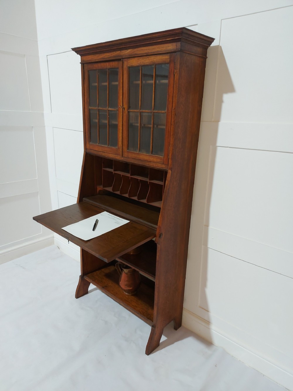 An Arts and Crafts oak bureau bookcase featuring a two-door glazed upper cabinet, a fall-front writing flap with internal pigeonholes, and lower open shelving. View II