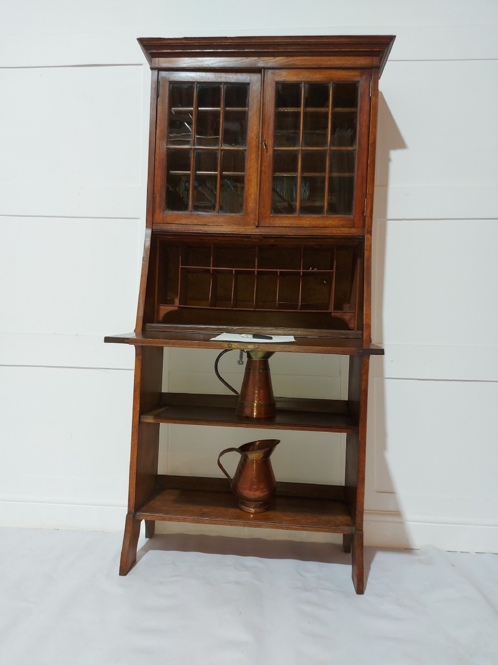 An Arts and Crafts oak bureau bookcase featuring a two-door glazed upper cabinet, a fall-front writing flap with internal pigeonholes, and lower open shelving. View V