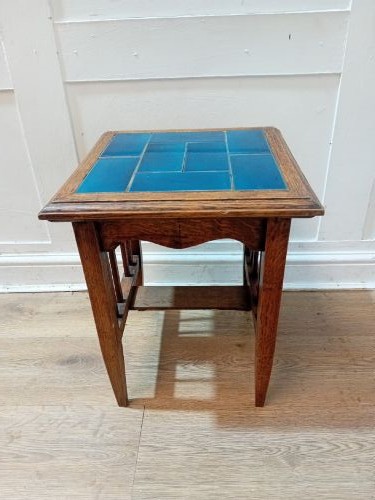 An Arts and Crafts style oak side table featuring a square top inset with vibrant blue ceramic tiles, straight tapered legs, and a lower stretcher shelf.
