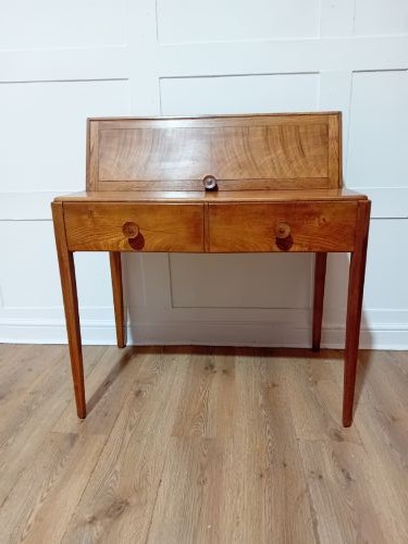 An Arts and Crafts Cotswold School wooden writing bureau featuring a fall-front upper section, two lower drawers with turned wooden knobs, and slender tapered legs. View V