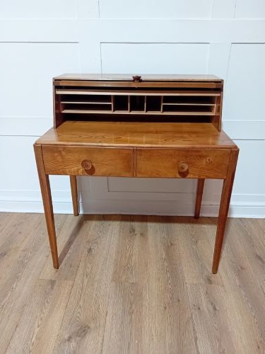 An Arts and Crafts Cotswold School wooden writing bureau featuring a fall-front upper section, two lower drawers with turned wooden knobs, and slender tapered legs. View VI
