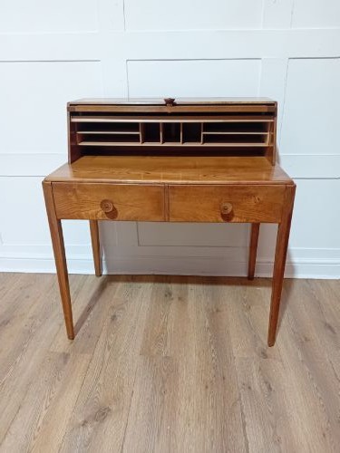 An Arts and Crafts Cotswold School wooden writing bureau featuring a fall-front upper section, two lower drawers with turned wooden knobs, and slender tapered legs. View IX