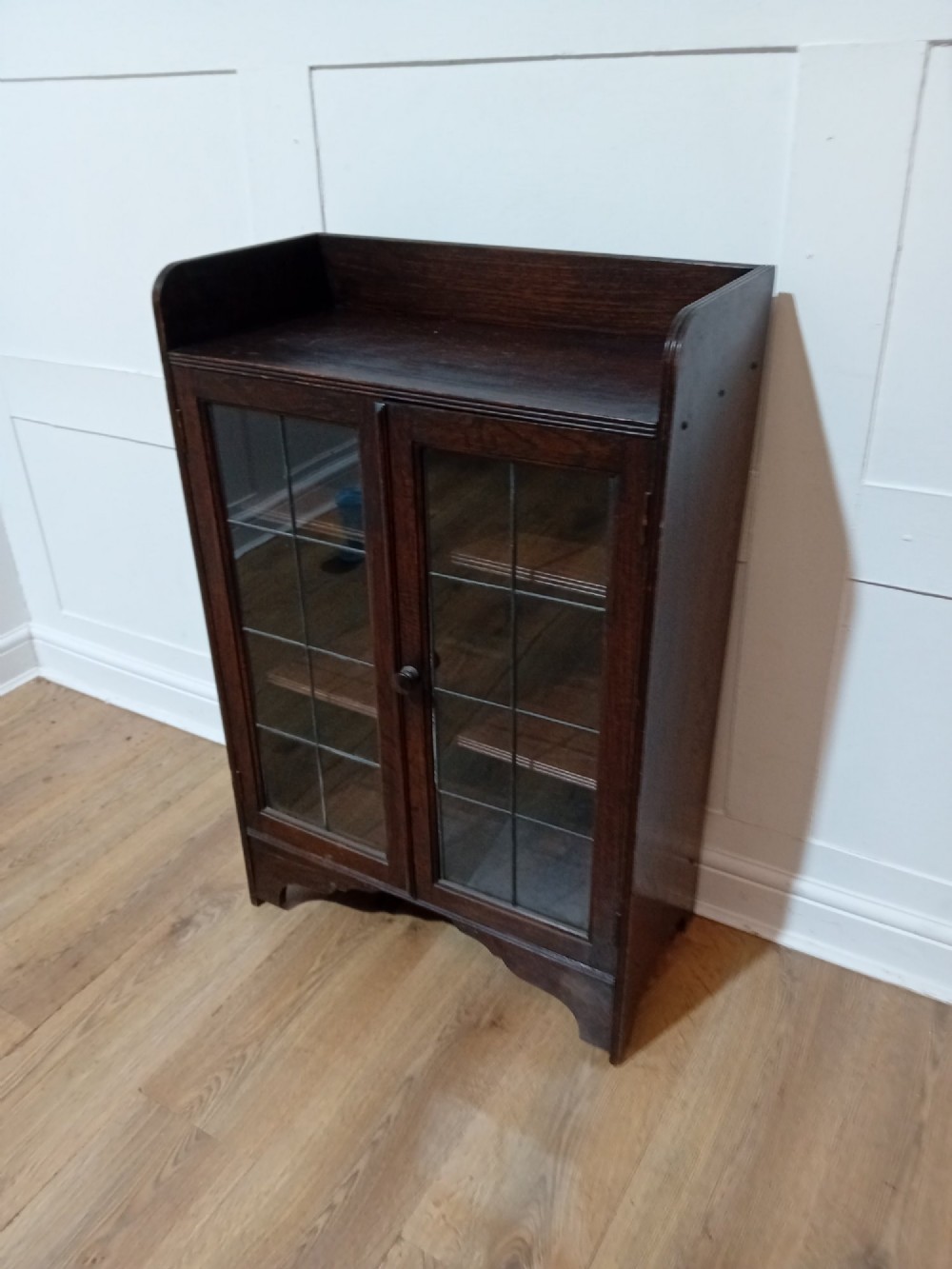 A dark oak Arts and Crafts dwarf glazed cabinet featuring twin leaded glass doors and a gallery top upstand.