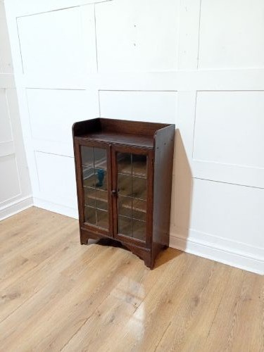 A dark oak Arts and Crafts dwarf glazed cabinet featuring twin leaded glass doors and a gallery top upstand. View II