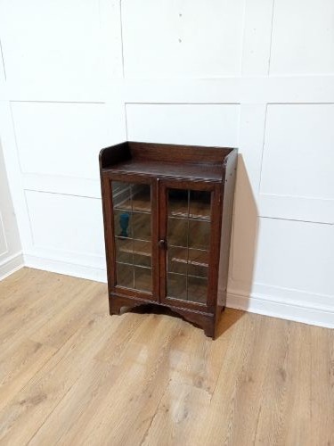 A dark oak Arts and Crafts dwarf glazed cabinet featuring twin leaded glass doors and a gallery top upstand. View III