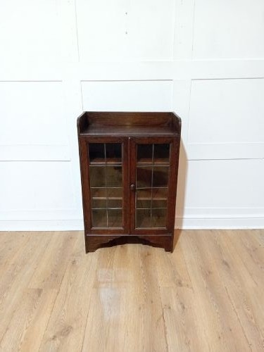 A dark oak Arts and Crafts dwarf glazed cabinet featuring twin leaded glass doors and a gallery top upstand. View IV