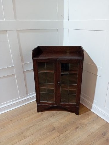 A dark oak Arts and Crafts dwarf glazed cabinet featuring twin leaded glass doors and a gallery top upstand. View V