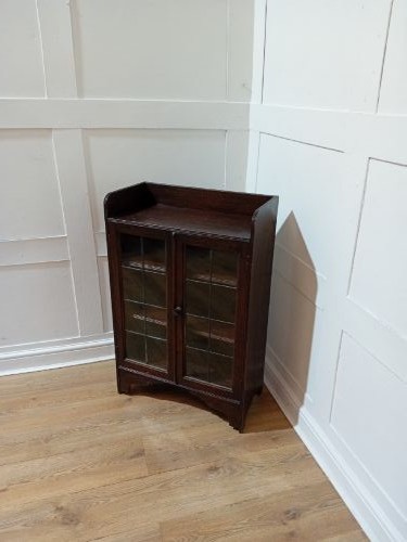 A dark oak Arts and Crafts dwarf glazed cabinet featuring twin leaded glass doors and a gallery top upstand. View VI