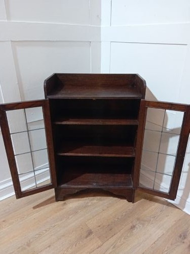 A dark oak Arts and Crafts dwarf glazed cabinet featuring twin leaded glass doors and a gallery top upstand. View IX