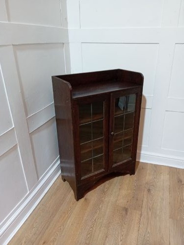 A dark oak Arts and Crafts dwarf glazed cabinet featuring twin leaded glass doors and a gallery top upstand. View VIII