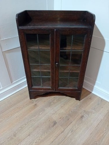 A dark oak Arts and Crafts dwarf glazed cabinet featuring twin leaded glass doors and a gallery top upstand. View XV