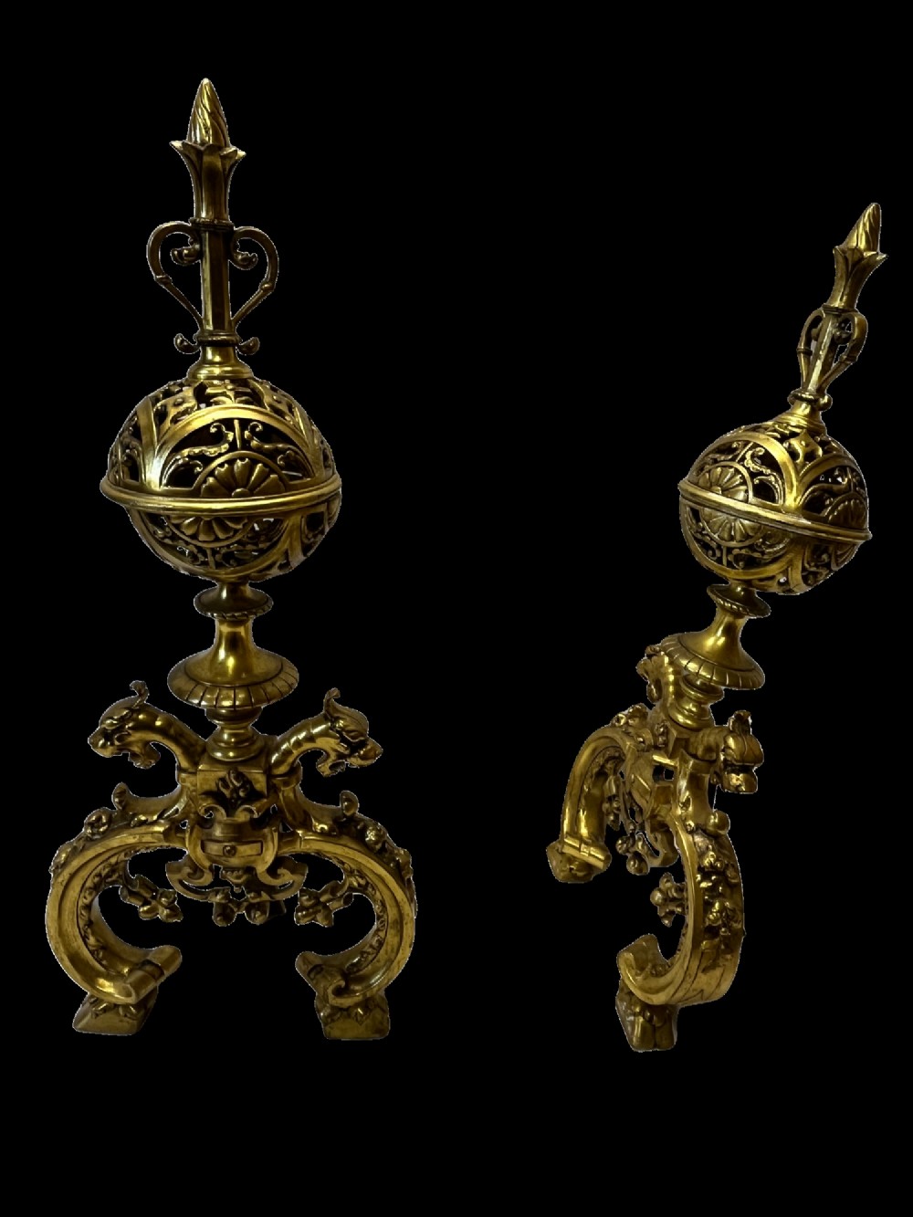 A pair of cast brass French fire dogs featuring pierced spherical tops, flame finials, and scrolling bases with carved animal heads.
