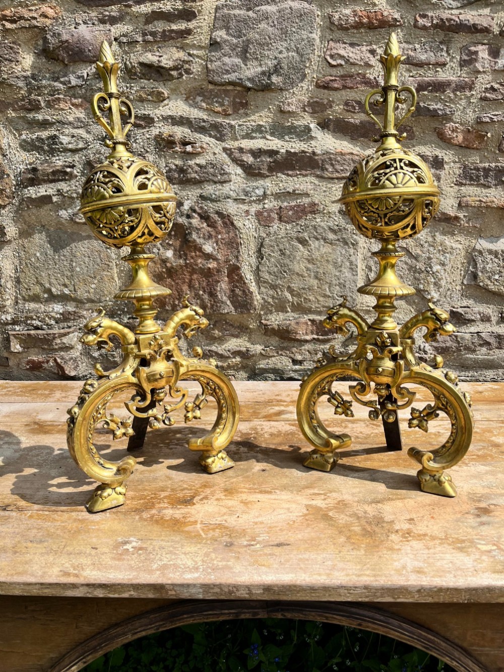 A pair of cast brass French fire dogs featuring pierced spherical tops, flame finials, and scrolling bases with carved animal heads. View II