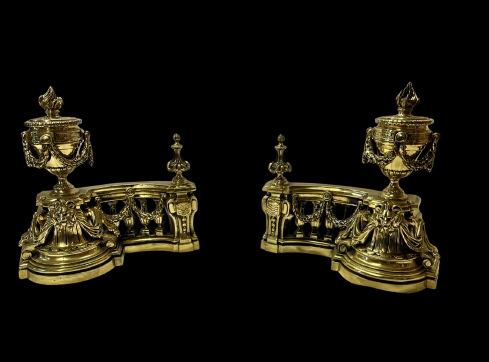 A pair of ornate brass fire dogs in the Louis XVI style, featuring draped flaming urns atop balustraded bases.