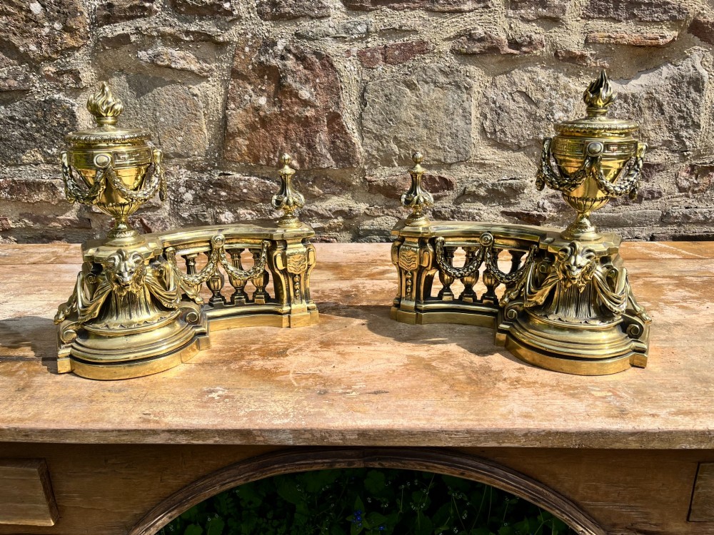 A pair of ornate brass fire dogs in the Louis XVI style, featuring draped flaming urns atop balustraded bases. View IV