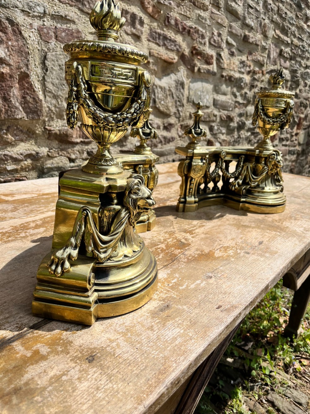 A pair of ornate brass fire dogs in the Louis XVI style, featuring draped flaming urns atop balustraded bases. View V