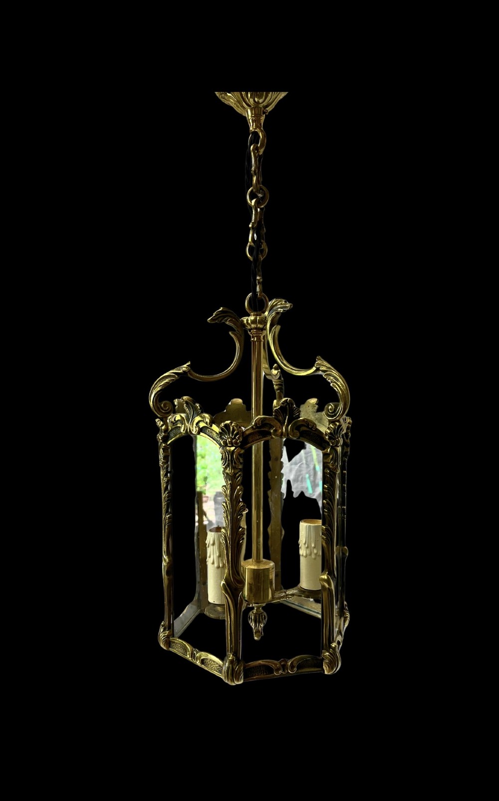 An ornate French style brass hanging hall lantern featuring glazed panels, acanthus scroll details, and three interior faux candle light sockets.