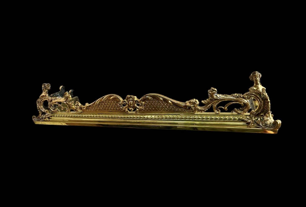 An ornate brass and bronze fireplace fender featuring Rococo scrollwork, lattice panels, and female figural end ornaments.