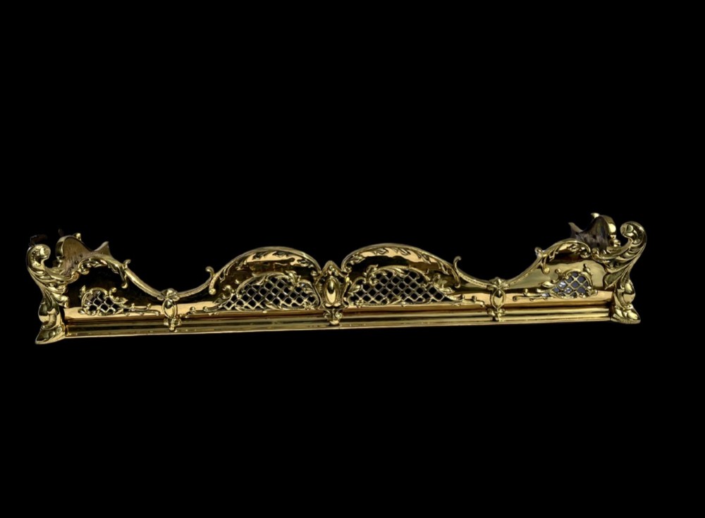 A late Victorian polished brass fireplace fender featuring pierced lattice panels, scrolling Rococo cresting, and shaped corner mounts against a dark background.