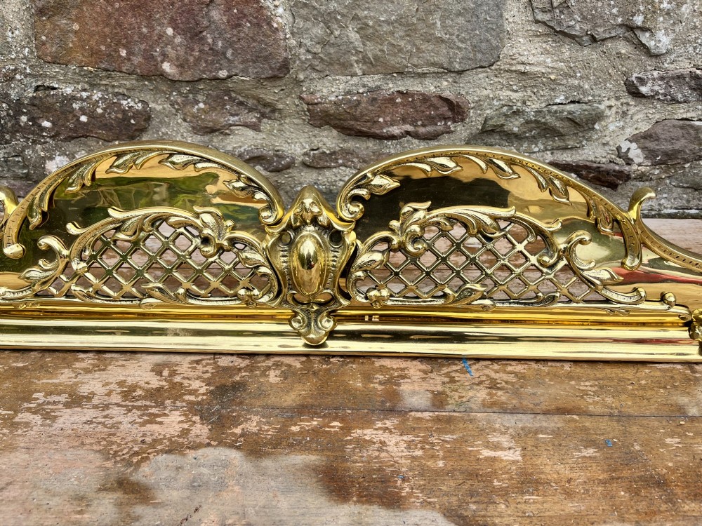 A late Victorian polished brass fireplace fender featuring pierced lattice panels, scrolling Rococo cresting, and shaped corner mounts against a dark background. View IV