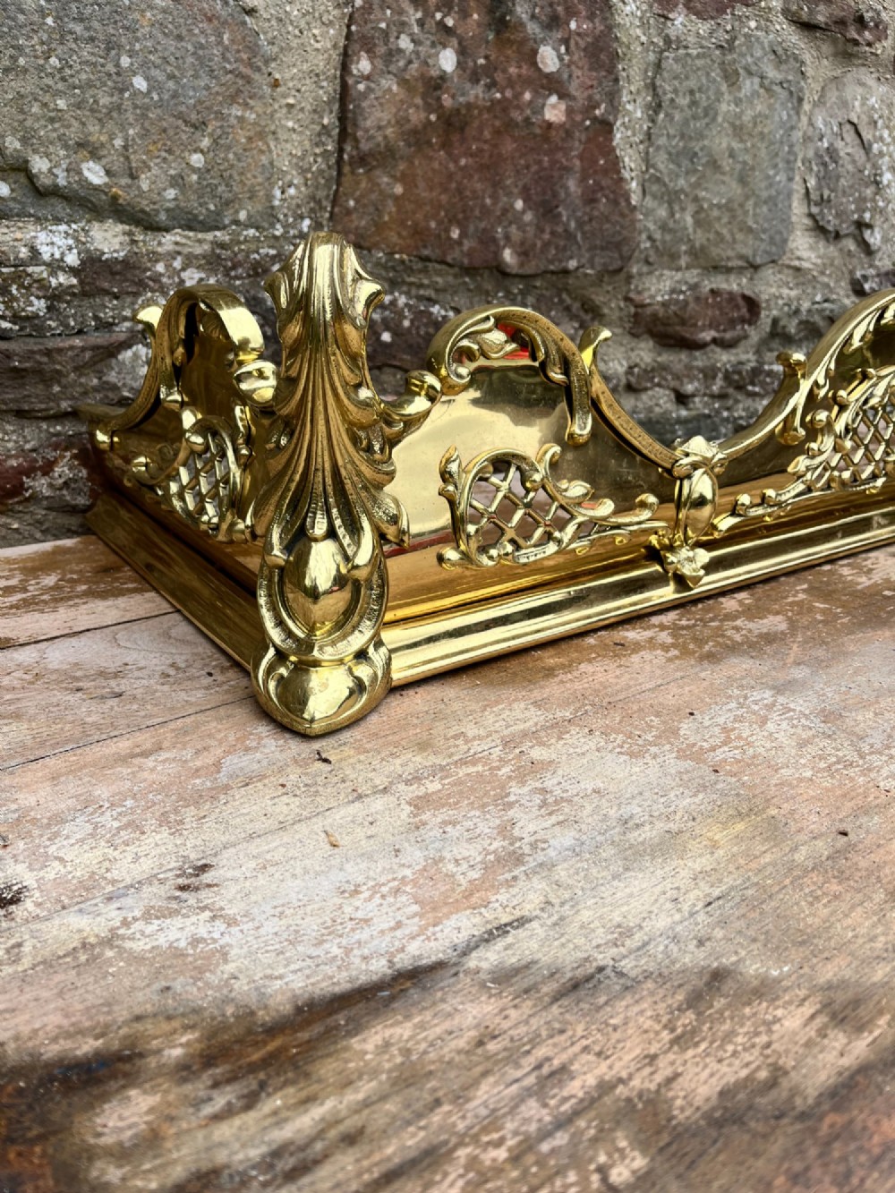 A late Victorian polished brass fireplace fender featuring pierced lattice panels, scrolling Rococo cresting, and shaped corner mounts against a dark background. View V