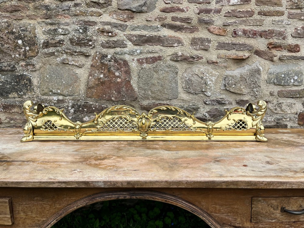A late Victorian polished brass fireplace fender featuring pierced lattice panels, scrolling Rococo cresting, and shaped corner mounts against a dark background. View VI