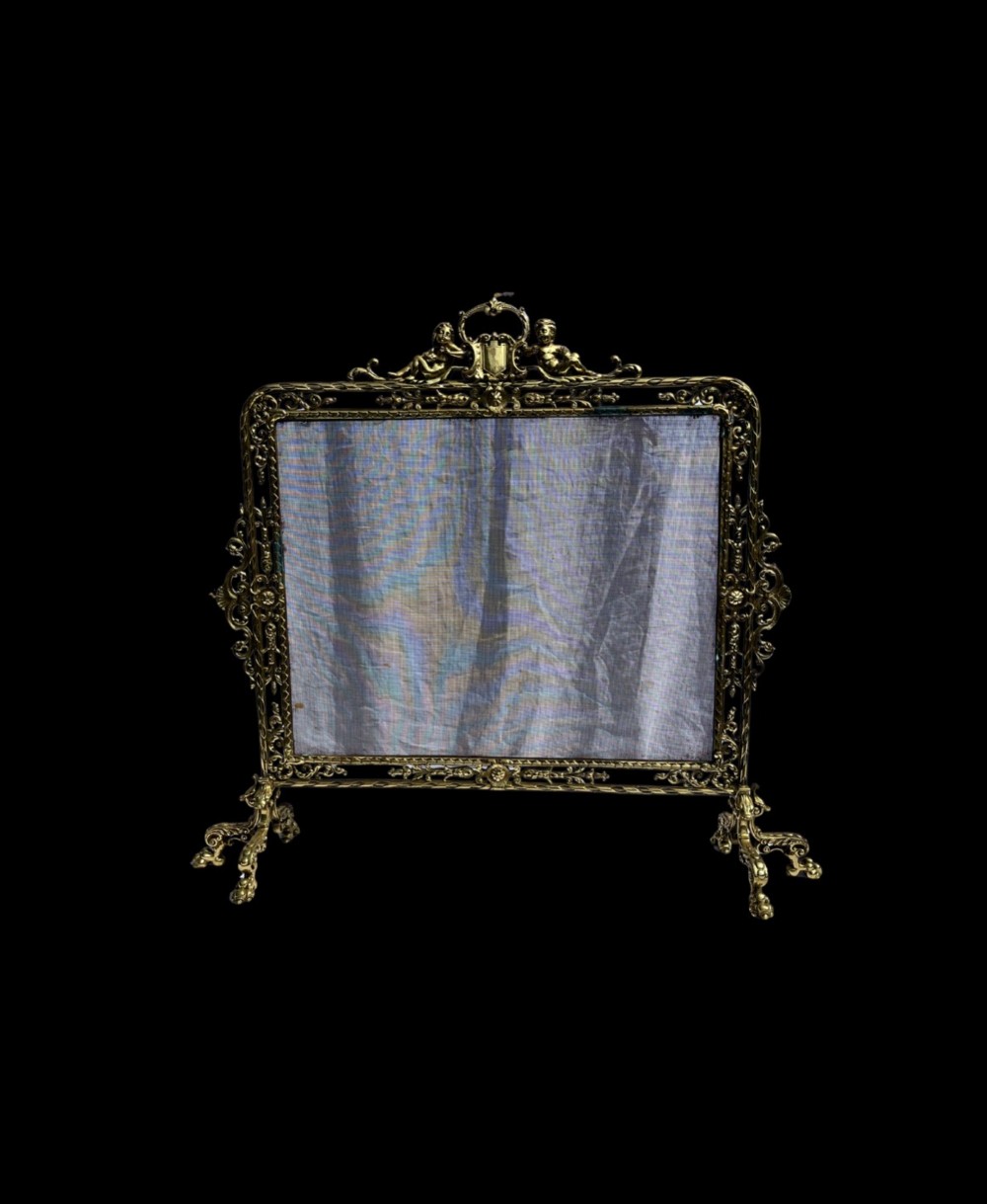 An ornate cast brass fire screen featuring cherub cresting, foliate side scrollwork, fine wire mesh insert, and four decorative feet.