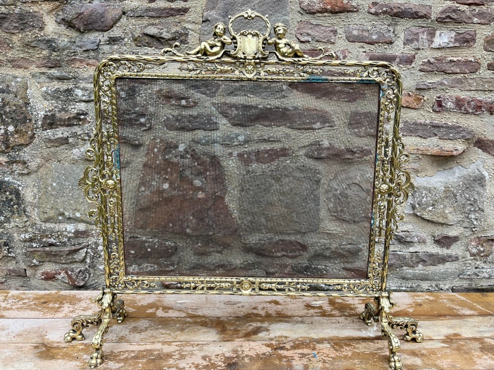 An ornate cast brass fire screen featuring cherub cresting, foliate side scrollwork, fine wire mesh insert, and four decorative feet. View III