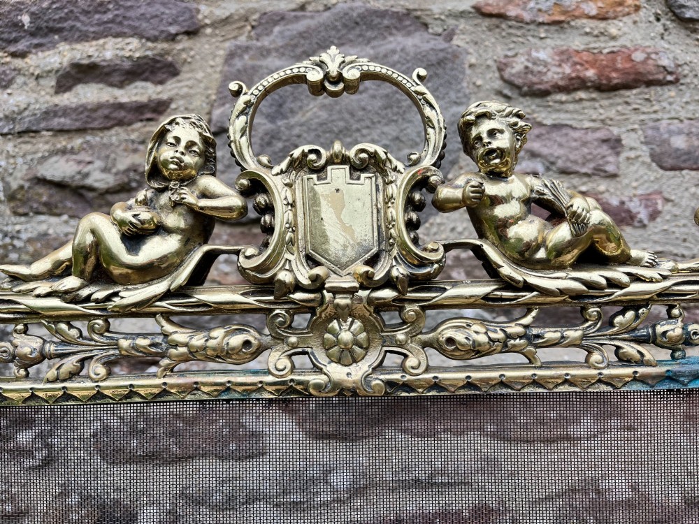 An ornate cast brass fire screen featuring cherub cresting, foliate side scrollwork, fine wire mesh insert, and four decorative feet. View V