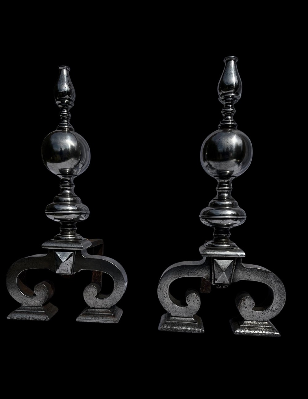 A pair of polished iron fire dogs featuring turned spherical finials and scrolled feet. View II