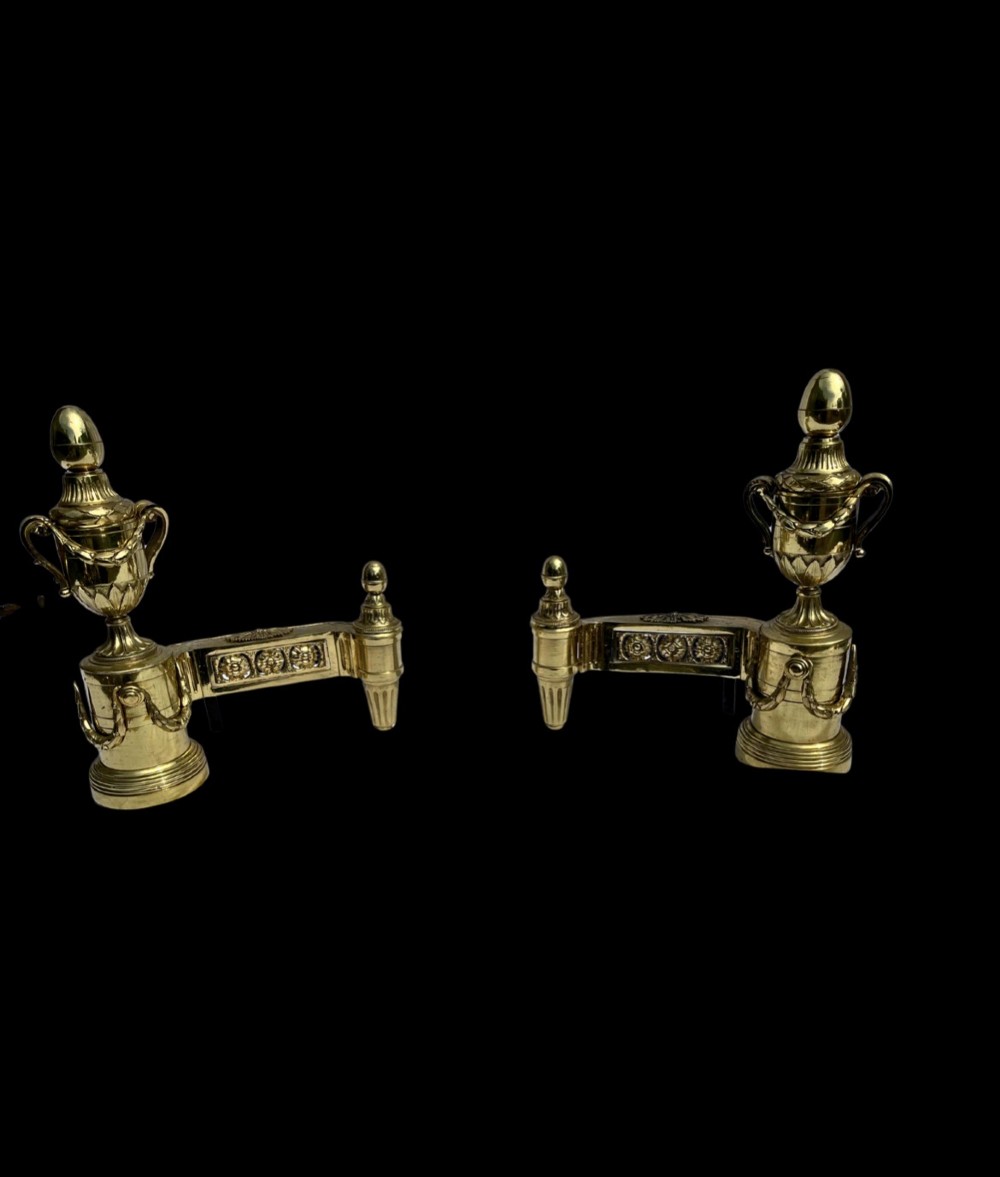 A pair of Neoclassical polished brass fire dogs featuring classical urn finials and garland swags. View II