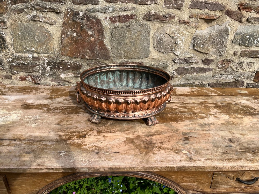 An antique circular copper jardiniere featuring gadrooned repousse patterns, side lion-mask ring handles, and three claw feet. View IV