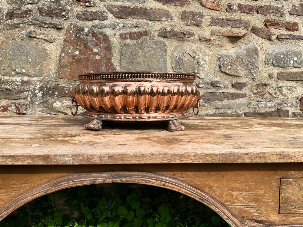 An antique circular copper jardiniere featuring gadrooned repousse patterns, side lion-mask ring handles, and three claw feet. View V